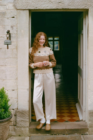 Woman standing in a doorway wearing a patterned sweater and white pants.