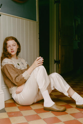 Woman sitting on a checkered floor holding a cup, wearing a patterned sweater and white pants.