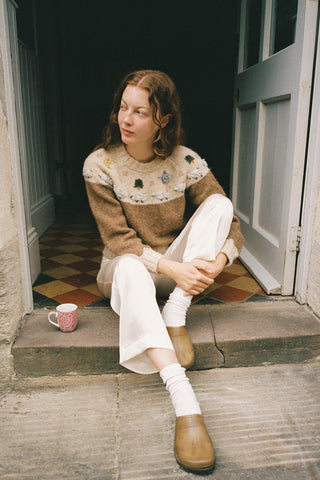 Woman sitting on a step wearing a patterned sweater and white pants, with a cup beside her.