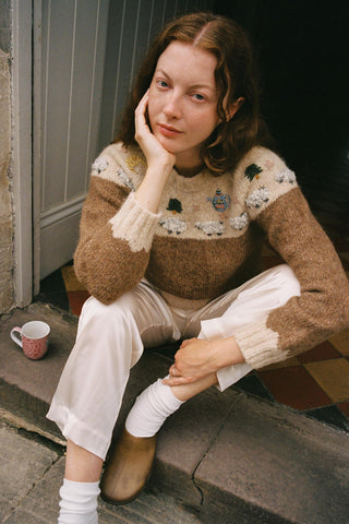 Woman sitting on steps wearing a patterned sweater and white pants, holding a mug.