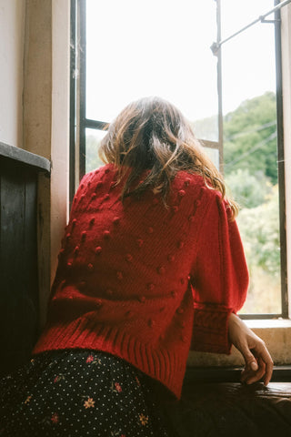 Person wearing a red sweater sitting by a window with a view of greenery.