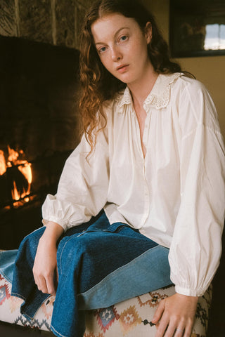 Woman in a white blouse and blue skirt sitting in front of a fireplace.
