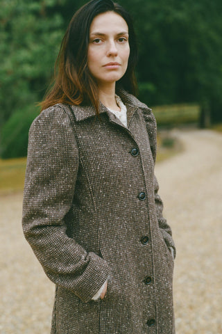 Woman wearing a long brown coat standing outdoors with greenery in the background