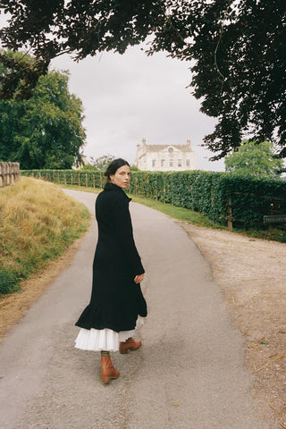 Woman in a black dress walking on a path with greenery and a house in the background
