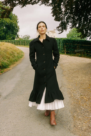 Woman in a long black coat and white dress standing on a road with greenery and a house in the background.