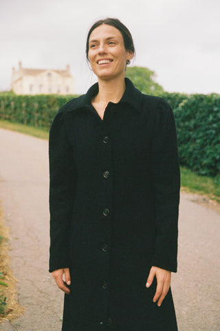 Person wearing a black coat standing outdoors with a building and greenery in the background