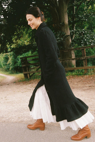 Woman wearing a long black coat over a white dress with brown shoes, standing outdoors.