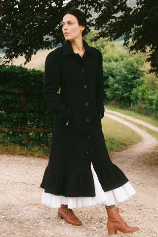 Woman wearing a long black coat with a white skirt standing on a gravel path with greenery in the background