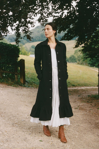 Woman wearing a long black coat over a white dress, standing on a path with greenery in the background.
