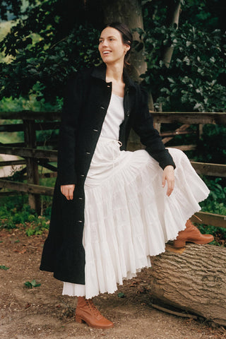 Woman in a black coat and white dress standing outdoors near a wooden fence.