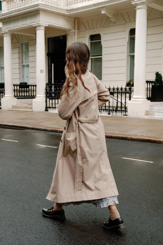 Person wearing a beige trench coat walking on a street in front of a building.