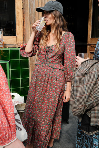 Woman in a patterned dress and cap drinking from a glass in an outdoor setting.