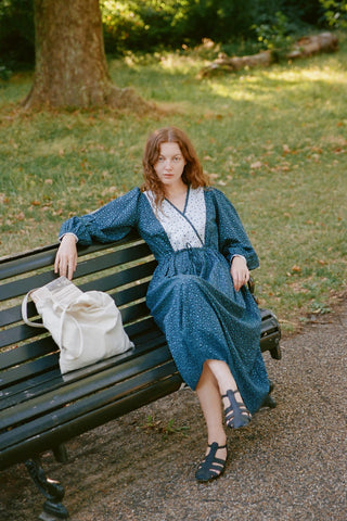Woman in a blue dress sitting on a bench in a park with a white bag beside her.