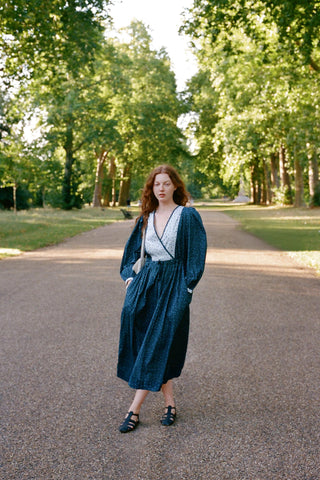 Woman in a blue dress with white polka dot trim and a deep v neck and tote bag on her shoulder standing on a gravel path