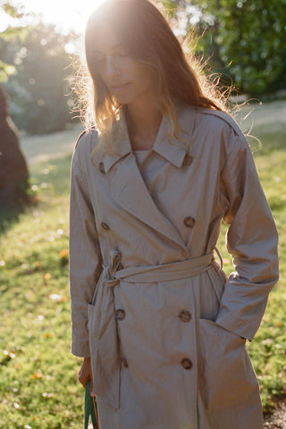 Woman wearing a beige trench coat standing in a grassy area with sunlight filtering through.