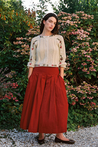 Woman wearing baggy red pants and a beige cardigan with greenery in the background 