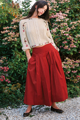 Woman wearing baggy red pants and a beige cardigan with greenery in the background 