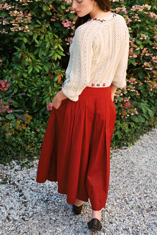 Woman facing away from camera wearing baggy red pants and a beige cardigan with greenery in the background 