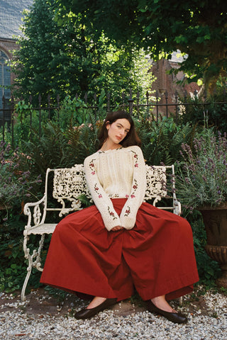 Woman sitting on a bench wearing baggy red pants and a beige cardigan with greenery in the background 