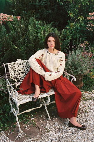 Woman sitting on a bench wearing baggy red pants and a beige cardigan with greenery in the background 