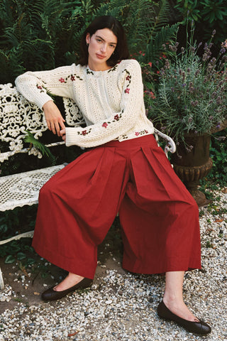 Woman sitting on a bench wearing baggy red pants and a beige cardigan with greenery in the background 