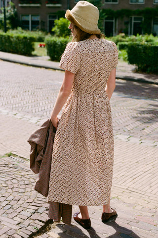 Women facing away from camera wearing a beige dress with a cozy hat holding a plaid item standing on cobblestones 