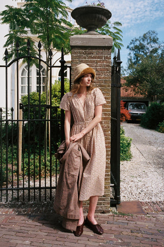 Women wearing a beige dress with a cozy hat holding a plaid item against brick