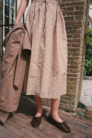 The bottom of a beige dress and brown shows with a plaid jacket standing on bricks