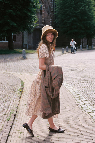A women walking across a street wearing a beige dress with a cozy hat holding a plaid jacket