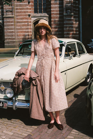 Women wearing a beige dress with a cozy hat holding a plaid item leaning against a vintage car