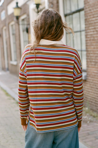 A person viewed from behind wearing a long-sleeve collared knit sweater with horizontal stripes in brown, red, blue, cream, and tan, paired with blue jeans, standing on a cobblestone street.
