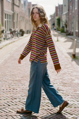 A woman walking on a sunlit brick street wearing a long-sleeve collared knit top with horizontal multicolor stripes in red, yellow, green, blue, and brown, paired with wide-leg blue jeans and brown loafers.