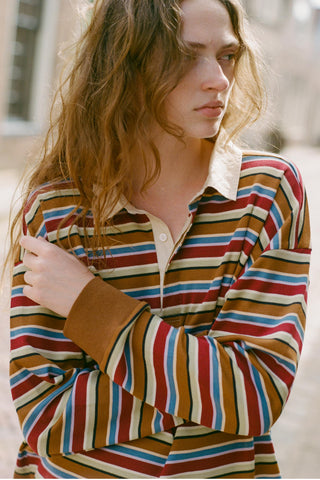 A woman wearing a long-sleeve collared knit top with horizontal stripes in red, blue, brown, and cream, photographed outdoors with a softly blurred background.