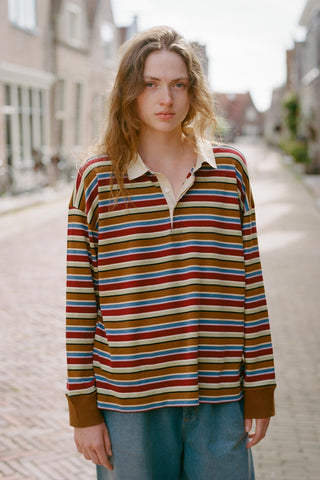 A young woman stands on a cobblestone street wearing a long-sleeve collared knit top with horizontal stripes in brown, red, blue, beige, and white, paired with light blue jeans.