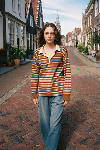 A woman stands on a brick street wearing a long-sleeve striped polo shirt with blue jeans. The shirt features horizontal stripes in brown, orange, red, and yellow over a cream base, with solid brown cuffs.