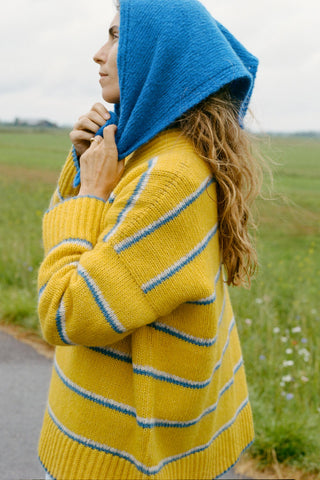 A woman wearing a bright yellow knit sweater with blue and white horizontal stripes, paired with a matching blue knit hood, standing outdoors in a grassy field.