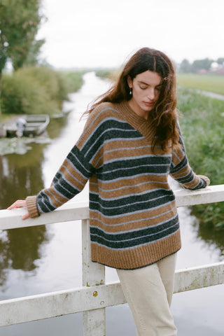 A woman wearing a cozy, oversized striped knit sweater leans against a white wooden bridge railing next to a calm canal, with green fields in the background.