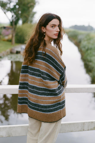 A woman wearing a cozy, oversized striped knit sweater leans against a white wooden bridge railing