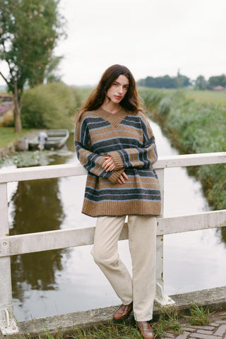 A woman wearing a cozy, oversized striped knit sweater leans against a white wooden bridge railing next to a calm canal
