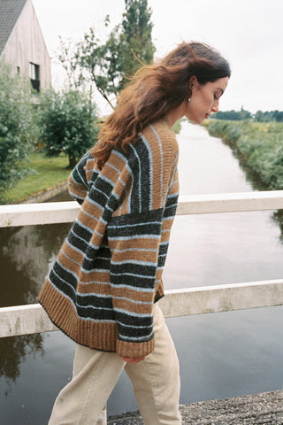 A woman wearing a chunky knit striped sweater in brown, beige, and gray, standing on a bridge overlooking a canal.