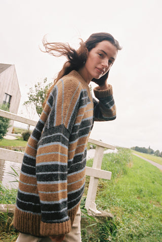 A woman outdoors wearing an oversized striped knit sweater with horizontal bands of brown, black, gray, and light blue, standing near a white fence in a grassy field.
