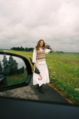 Woman wearing a colorful vest over a white shirt and skirt on a street with a green field behind her