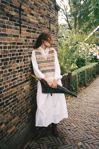 Woman wearing a colorful vest over a white shirt and skirt leaning on bricks