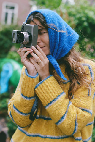 A woman wearing a bright yellow knit sweater with blue and white horizontal stripes, paired with a matching blue knit hood holding a film camera

