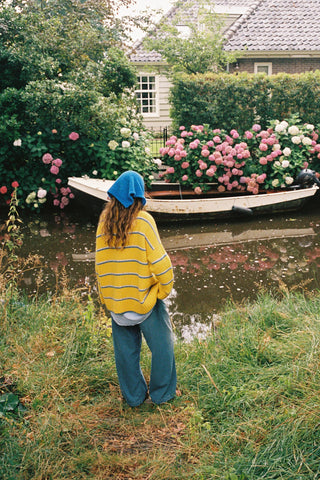 A woman wearing a bright yellow knit sweater with blue and white horizontal stripes, paired with a matching blue knit hood, standing outdoors in a grassy field 
looking at water