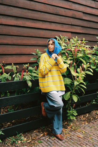 A person wearing a yellow and white striped knit sweater, blue jeans, and a blue headscarf leans against a dark wooden fence next to green plants with red flowers.