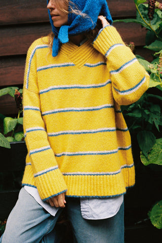A woman wearing a bright yellow knit sweater with blue and white horizontal stripes, paired with a matching blue knit hood and jeans

