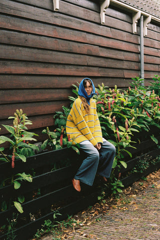 A woman wearing a bright yellow knit sweater with blue and white horizontal stripes, paired with a matching blue knit hood, sitting on a fence
