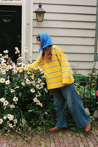 A woman wearing a bright yellow knit sweater with blue and white horizontal stripes, paired with a matching blue knit hood, reaching for flowers
