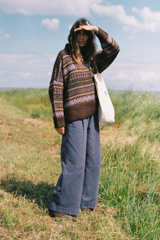 Woman wearing a patterned oversized sweater in a field with blue baggy pants and a bag on her shoulder